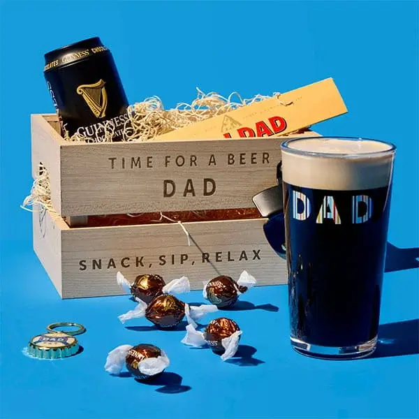 Fathers Day gift arrangement on a blue background featuring a wooden beer crate for dad, a pint of stout, chocolates, and a can of beer